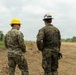 Two individuals wearing U.S. Marine Corps MARPAT digital camouflage uniforms and hard hats are standing on a cleared, earthen construction or engineering site. One wears a yellow hard hat and sunglasses, while the other wears a white hard hat with visible insignia. They appear to be overseeing or surveying a land-clearing or construction operation in a tropical or subtropical environment.