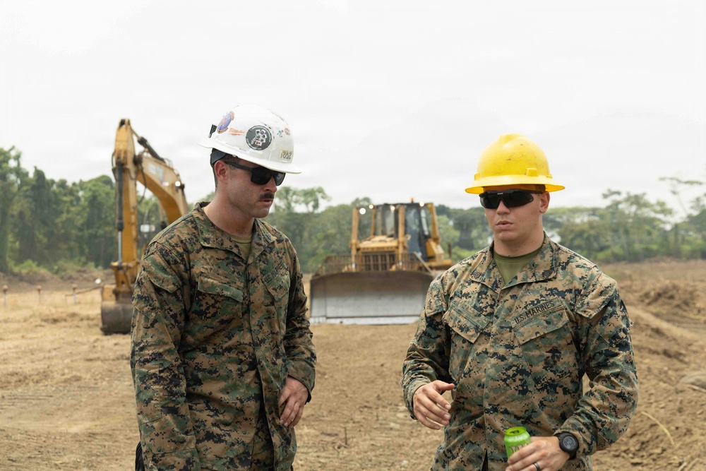 Two U.S. Marines in digital camouflage uniforms and hard hats are conversing at an active construction site. Heavy equipment including an excavator and a bulldozer are visible in the background on cleared dirt terrain. The setting appears to be a tropical or subtropical region based on the dense tree line visible in the background.