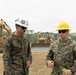 Two U.S. Marines in digital camouflage uniforms and hard hats are conversing at an active construction site. Heavy equipment including an excavator and a bulldozer are visible in the background on cleared dirt terrain. The setting appears to be a tropical or subtropical region based on the dense tree line visible in the background.