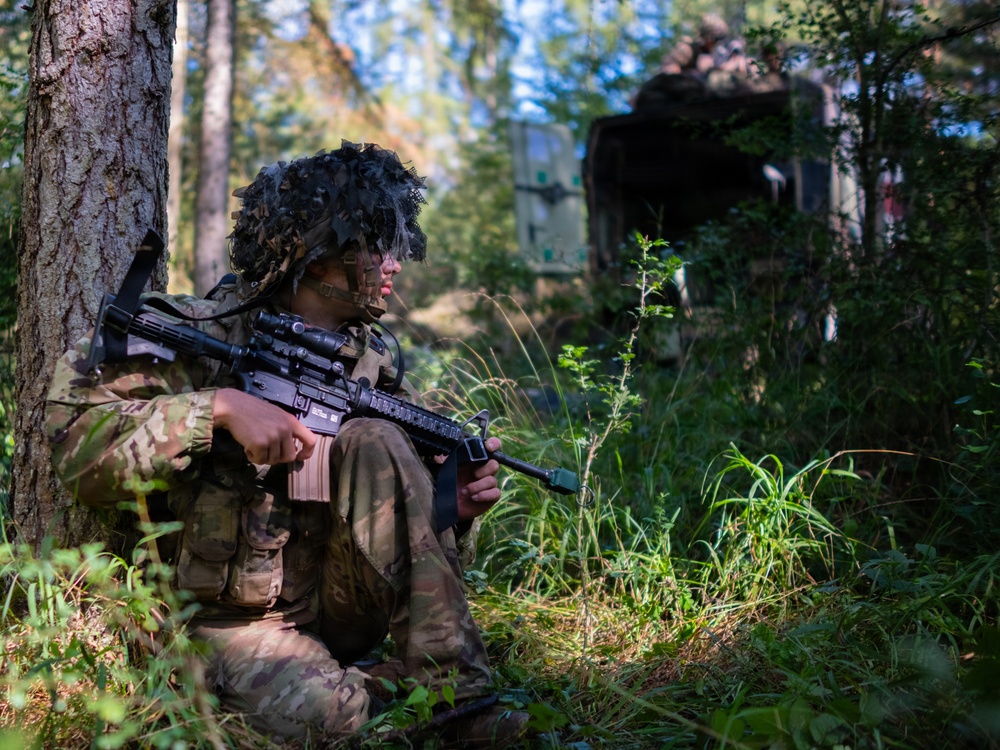 A soldier in full camouflage uniform and ghillie-style helmet cover is positioned against a tree in a forested area, holding an assault rifle in a security posture. The setting appears to be a military training exercise, with a tactical military vehicle visible in the background among the trees. The soldier appears alert and is scanning their surroundings.