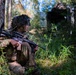 A soldier in full camouflage uniform and ghillie-style helmet cover is positioned against a tree in a forested area, holding an assault rifle in a security posture. The setting appears to be a military training exercise, with a tactical military vehicle visible in the background among the trees. The soldier appears alert and is scanning their surroundings.