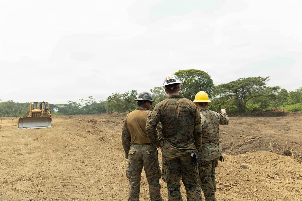 Three military personnel in camouflage uniforms wearing hard hats (gray, white, and yellow) are observing a cleared construction site. A large yellow bulldozer/dozer is visible in the background operating on bare earth. The scene appears to be a military engineering or construction operation in a tropical or subtropical environment.