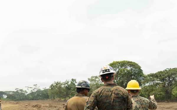 Three military personnel in camouflage uniforms wearing hard hats (gray, white, and yellow) are observing a cleared construction site. A large yellow bulldozer/dozer is visible in the background operating on bare earth. The scene appears to be a military engineering or construction operation in a tropical or subtropical environment.