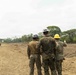 Three military personnel in camouflage uniforms wearing hard hats (gray, white, and yellow) are observing a cleared construction site. A large yellow bulldozer/dozer is visible in the background operating on bare earth. The scene appears to be a military engineering or construction operation in a tropical or subtropical environment.