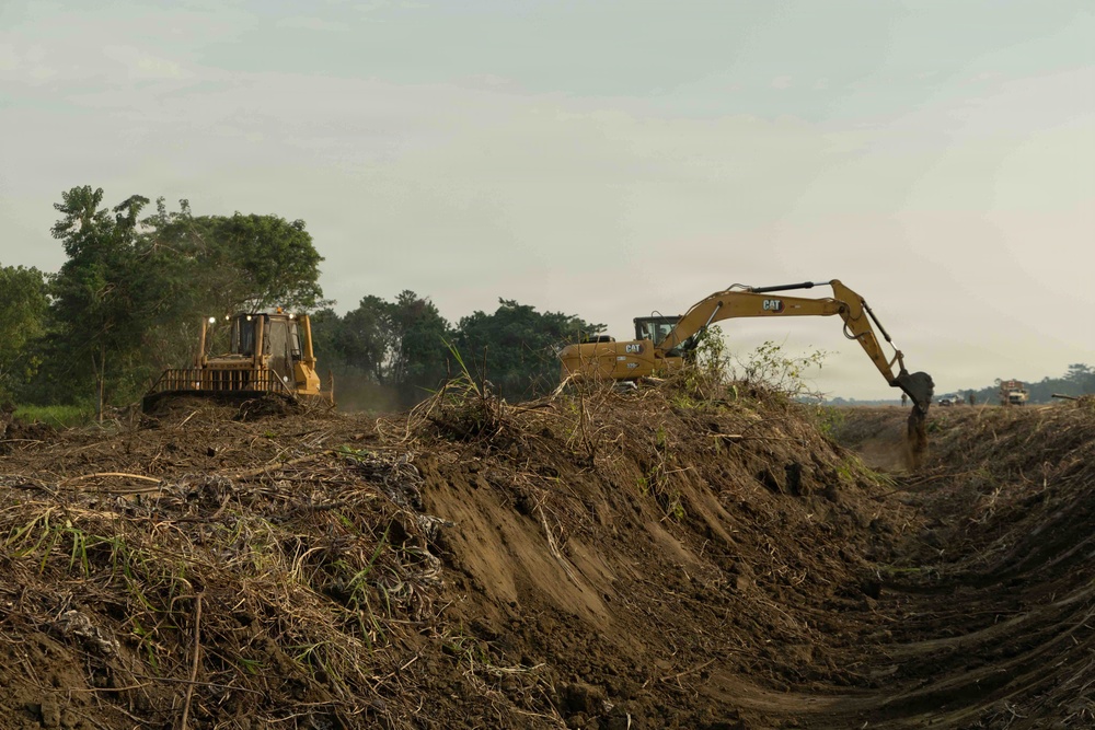 Two yellow Caterpillar (CAT) heavy construction machines are actively working on a large earthmoving operation in what appears to be a tropical or subtropical environment. A bulldozer on the left is pushing soil and debris while an excavator on the right is digging into a mounded earthen berm. The surrounding area shows cleared vegetation with dense trees visible in the background, suggesting land clearing or infrastructure development.