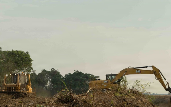 Two yellow Caterpillar (CAT) heavy construction machines are actively working on a large earthmoving operation in what appears to be a tropical or subtropical environment. A bulldozer on the left is pushing soil and debris while an excavator on the right is digging into a mounded earthen berm. The surrounding area shows cleared vegetation with dense trees visible in the background, suggesting land clearing or infrastructure development.