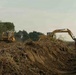 Two yellow Caterpillar (CAT) heavy construction machines are actively working on a large earthmoving operation in what appears to be a tropical or subtropical environment. A bulldozer on the left is pushing soil and debris while an excavator on the right is digging into a mounded earthen berm. The surrounding area shows cleared vegetation with dense trees visible in the background, suggesting land clearing or infrastructure development.