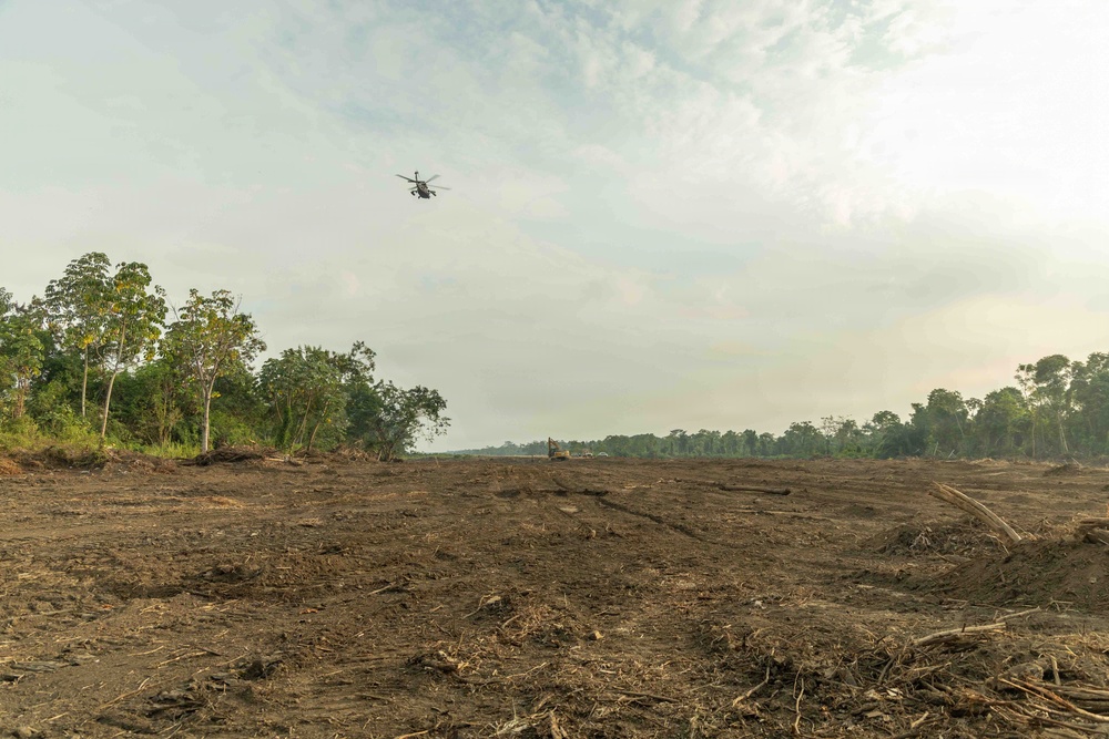 The image shows a large area of recently cleared forest land, with bare, churned soil and debris covering the foreground. A helicopter is visible flying low over the cleared area, and what appears to be heavy machinery is visible in the mid-ground. Intact forest tree lines remain visible on the left, right, and background edges of the cleared zone.