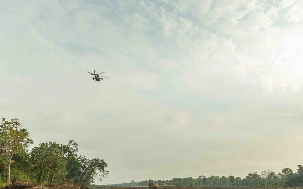 The image shows a large area of recently cleared forest land, with bare, churned soil and debris covering the foreground. A helicopter is visible flying low over the cleared area, and what appears to be heavy machinery is visible in the mid-ground. Intact forest tree lines remain visible on the left, right, and background edges of the cleared zone.