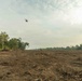 The image shows a large area of recently cleared forest land, with bare, churned soil and debris covering the foreground. A helicopter is visible flying low over the cleared area, and what appears to be heavy machinery is visible in the mid-ground. Intact forest tree lines remain visible on the left, right, and background edges of the cleared zone.