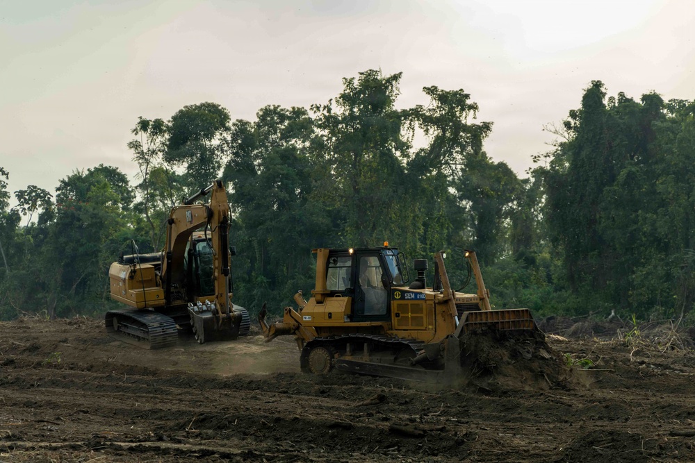 Two large yellow construction machines are actively clearing land at the edge of a dense tropical or subtropical forest. A tracked excavator and a bulldozer (branded 'SEM 816D') are operating on freshly cleared, bare earth. The background shows a wall of green trees, indicating active deforestation or land clearing activity.