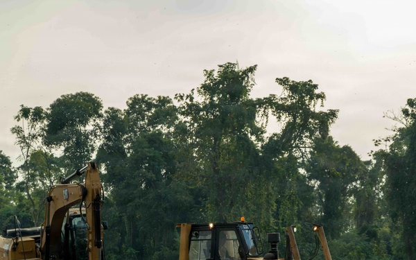 Two large yellow construction machines are actively clearing land at the edge of a dense tropical or subtropical forest. A tracked excavator and a bulldozer (branded 'SEM 816D') are operating on freshly cleared, bare earth. The background shows a wall of green trees, indicating active deforestation or land clearing activity.