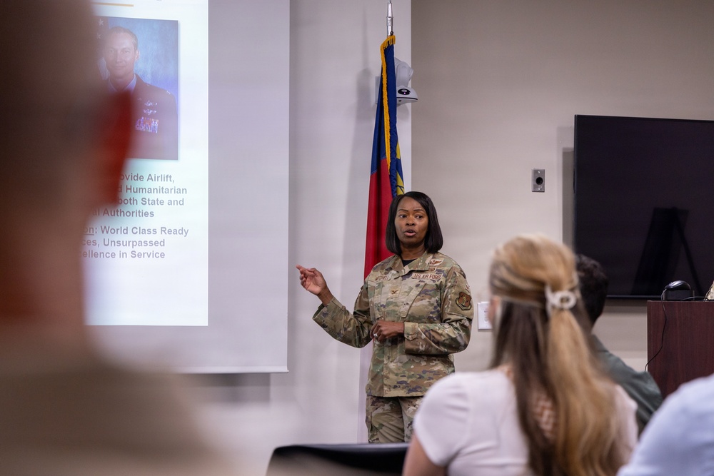 A US Air Force service member in OCP (Operational Camouflage Pattern) uniform is delivering a briefing or presentation to a small audience in a conference room. A projection screen displays a slide with text referencing 'Airlift,' 'Humanitarian,' and 'World Class Ready' alongside a portrait photo of a military officer. A state flag, identifiable as the North Carolina flag, is visible in the background.