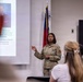 A US Air Force service member in OCP (Operational Camouflage Pattern) uniform is delivering a briefing or presentation to a small audience in a conference room. A projection screen displays a slide with text referencing 'Airlift,' 'Humanitarian,' and 'World Class Ready' alongside a portrait photo of a military officer. A state flag, identifiable as the North Carolina flag, is visible in the background.