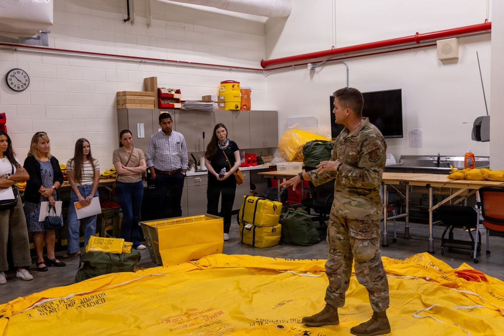 A military service member in camouflage uniform is conducting a briefing or demonstration for a group of civilians in what appears to be a military equipment storage or training facility. Large yellow equipment items, including what appears to be an inflatable or fabric material spread on the floor, are the focus of the presentation. The group of approximately six civilians is attentively observing the demonstration.