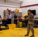 A military service member in camouflage uniform is conducting a briefing or demonstration for a group of civilians in what appears to be a military equipment storage or training facility. Large yellow equipment items, including what appears to be an inflatable or fabric material spread on the floor, are the focus of the presentation. The group of approximately six civilians is attentively observing the demonstration.