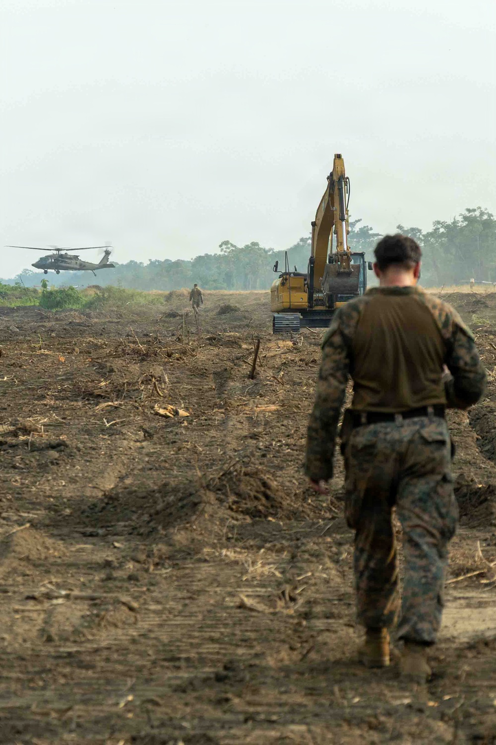 Military personnel in camouflage uniforms are walking across a cleared, recently graded dirt field toward heavy construction equipment and a hovering helicopter. The scene appears to depict a military engineering or construction operation, likely airfield or landing zone preparation. A yellow excavator and what appears to be a UH-60 Black Hawk helicopter are visible in the background amid a hazy, tropical environment.