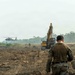 Military personnel in camouflage uniforms are walking across a cleared, recently graded dirt field toward heavy construction equipment and a hovering helicopter. The scene appears to depict a military engineering or construction operation, likely airfield or landing zone preparation. A yellow excavator and what appears to be a UH-60 Black Hawk helicopter are visible in the background amid a hazy, tropical environment.