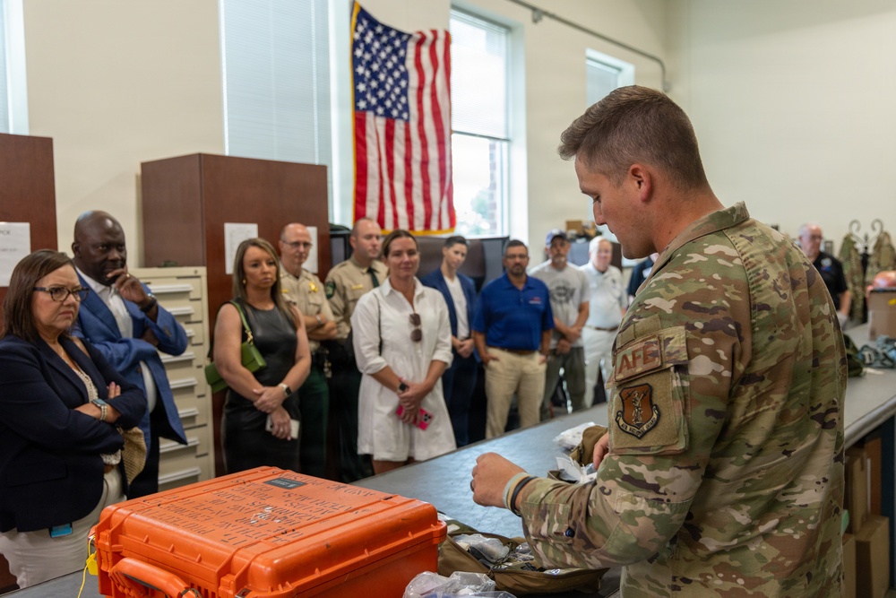 A U.S. Air Force service member in OCP (Operational Camouflage Pattern) uniform is conducting a briefing or demonstration for a group of civilian and law enforcement visitors in what appears to be a military facility. The presenter is standing at a table with an orange Pelican-style hard case and various equipment items. An American flag is displayed prominently on the wall in the background.