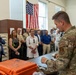 A U.S. Air Force service member in OCP (Operational Camouflage Pattern) uniform is conducting a briefing or demonstration for a group of civilian and law enforcement visitors in what appears to be a military facility. The presenter is standing at a table with an orange Pelican-style hard case and various equipment items. An American flag is displayed prominently on the wall in the background.