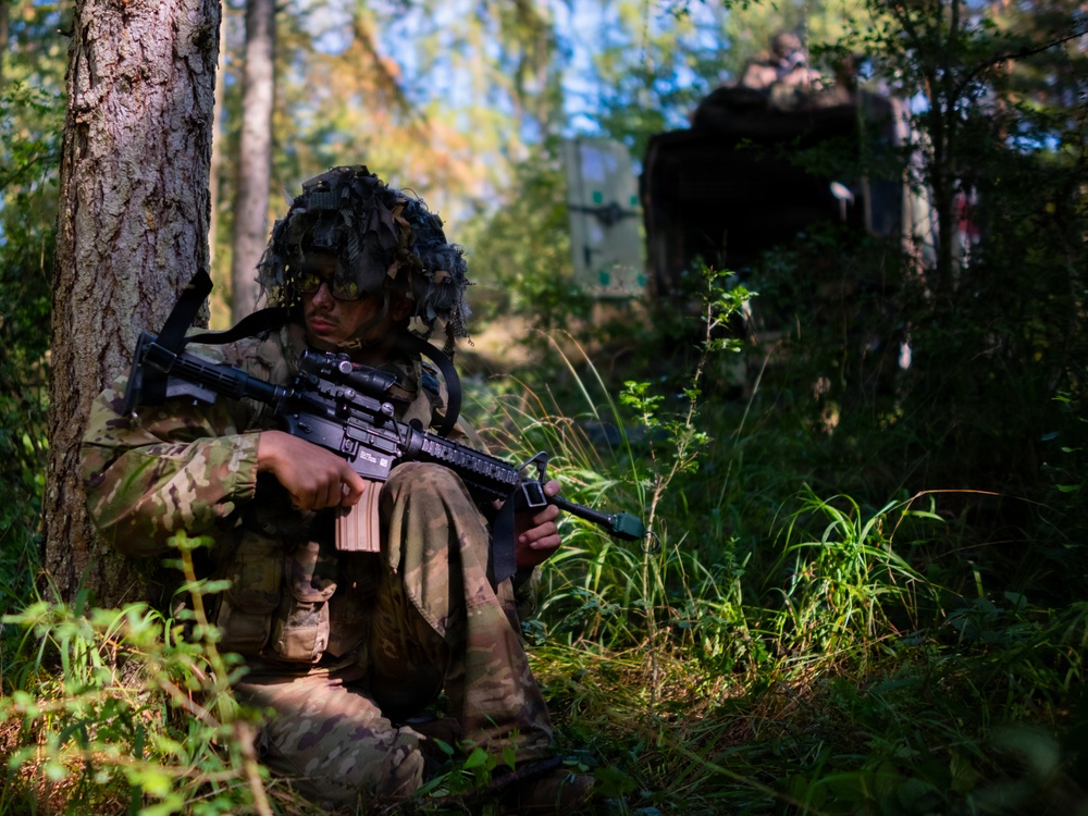 A soldier in full camouflage gear is seated against a tree in a forested environment, holding an assault rifle. The soldier is wearing a ghillie-style camouflaged helmet cover and MultiCam/OCP pattern uniform. A military vehicle or equipment is partially visible in the background through the trees.