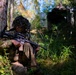 A soldier in full camouflage gear is seated against a tree in a forested environment, holding an assault rifle. The soldier is wearing a ghillie-style camouflaged helmet cover and MultiCam/OCP pattern uniform. A military vehicle or equipment is partially visible in the background through the trees.