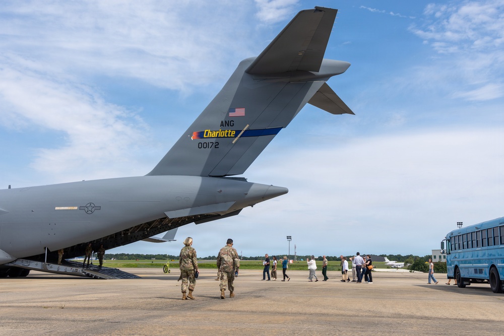 A large military transport aircraft, identified as belonging to the Air National Guard (ANG) unit based in Charlotte with tail number 00172, is parked on a tarmac. Two military personnel in camouflage uniforms are walking toward the aircraft's open rear cargo ramp, while a group of civilians stands nearby on the flight line. A light blue bus is visible on the right side of the image.