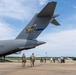 A large military transport aircraft, identified as belonging to the Air National Guard (ANG) unit based in Charlotte with tail number 00172, is parked on a tarmac. Two military personnel in camouflage uniforms are walking toward the aircraft's open rear cargo ramp, while a group of civilians stands nearby on the flight line. A light blue bus is visible on the right side of the image.