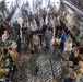 The image shows the interior of a large military cargo aircraft, likely a C-17 Globemaster III, filled with a mixed group of military personnel in camouflage uniforms and civilians in casual and business attire. The gathering appears to be an organized tour, briefing, or demonstration event, with military members presenting equipment and capabilities to civilian visitors. Medical and logistical equipment is visible along the sides of the aircraft's cargo bay.