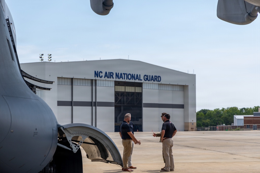 Two individuals in civilian attire (dark polo shirts and khaki pants) are conversing on an aircraft tarmac beside a large military aircraft. The setting is the NC Air National Guard facility, as indicated by the prominent signage on the hangar in the background. The scene appears to depict a professional discussion or briefing near the aircraft.