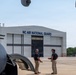 Two individuals in civilian attire (dark polo shirts and khaki pants) are conversing on an aircraft tarmac beside a large military aircraft. The setting is the NC Air National Guard facility, as indicated by the prominent signage on the hangar in the background. The scene appears to depict a professional discussion or briefing near the aircraft.