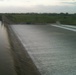 The image shows a large dam or spillway with water flowing over its face in a thin, uniform sheet. The structure appears to be a concrete gravity dam with a gated outlet visible at the base of the embankment. In the background, green vegetation, trees, and what appears to be a dome-shaped structure are visible on the horizon.