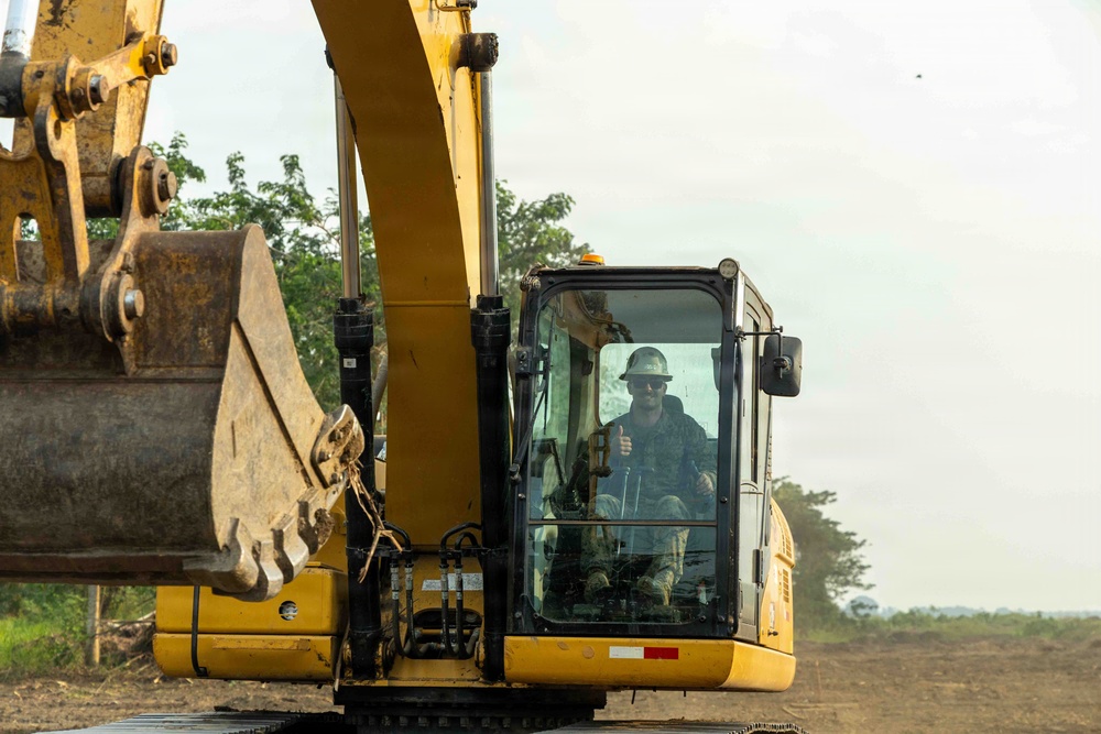 A operator wearing a white hard hat and military-style clothing is seated inside the cab of a large yellow excavator, giving a thumbs-up gesture toward the camera. The excavator's boom arm and bucket are prominently visible in the foreground. The setting appears to be an active earthmoving or construction site with cleared land and trees visible in the background.