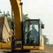 A operator wearing a white hard hat and military-style clothing is seated inside the cab of a large yellow excavator, giving a thumbs-up gesture toward the camera. The excavator's boom arm and bucket are prominently visible in the foreground. The setting appears to be an active earthmoving or construction site with cleared land and trees visible in the background.