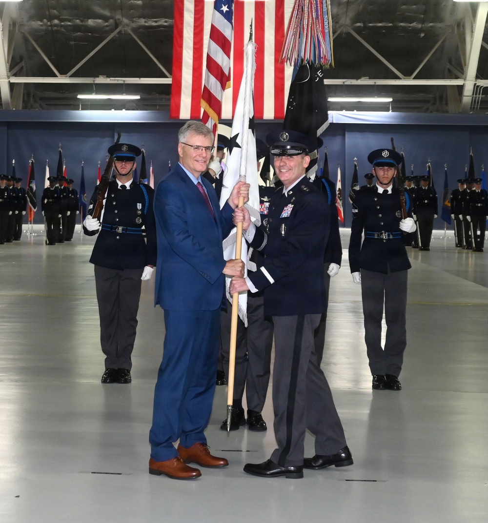 A formal military change of command or activation ceremony taking place inside a large hangar facility. Two central figures — a civilian in a blue suit and a uniformed military officer — are jointly holding a ceremonial guidon or unit flag. Honor guard members in dark dress uniforms are positioned in formation in the background alongside multiple flags including the American flag.