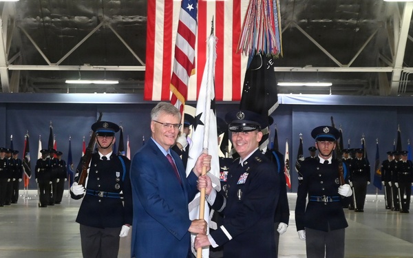A formal military change of command or activation ceremony taking place inside a large hangar facility. Two central figures — a civilian in a blue suit and a uniformed military officer — are jointly holding a ceremonial guidon or unit flag. Honor guard members in dark dress uniforms are positioned in formation in the background alongside multiple flags including the American flag.