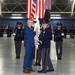 A formal military change of command or activation ceremony taking place inside a large hangar facility. Two central figures — a civilian in a blue suit and a uniformed military officer — are jointly holding a ceremonial guidon or unit flag. Honor guard members in dark dress uniforms are positioned in formation in the background alongside multiple flags including the American flag.