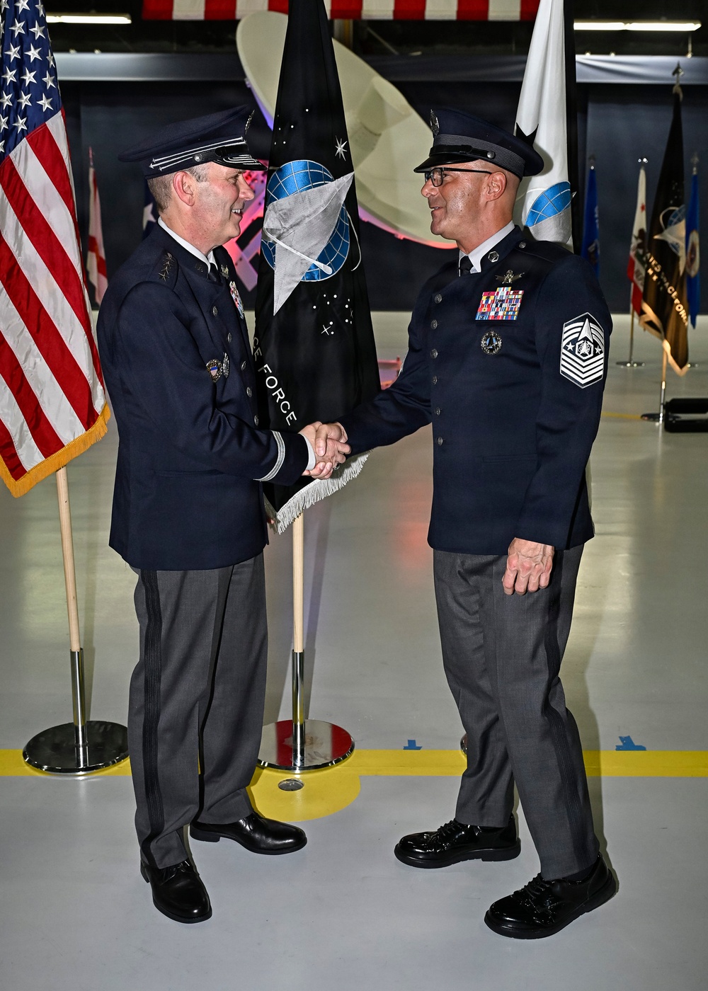Two military personnel in dress uniforms are shaking hands during a formal ceremony, with one holding a guidon/flag staff. The black flag in the background displays a globe emblem and the words 'FORCE,' consistent with United States Space Force insignia. The setting appears to be a military hangar decorated with multiple flags including the American flag.