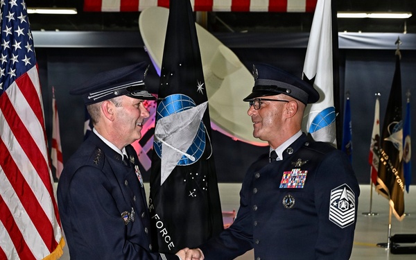 Two military personnel in dress uniforms are shaking hands during a formal ceremony, with one holding a guidon/flag staff. The black flag in the background displays a globe emblem and the words 'FORCE,' consistent with United States Space Force insignia. The setting appears to be a military hangar decorated with multiple flags including the American flag.