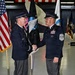 Two military personnel in dress uniforms are shaking hands during a formal ceremony, with one holding a guidon/flag staff. The black flag in the background displays a globe emblem and the words 'FORCE,' consistent with United States Space Force insignia. The setting appears to be a military hangar decorated with multiple flags including the American flag.