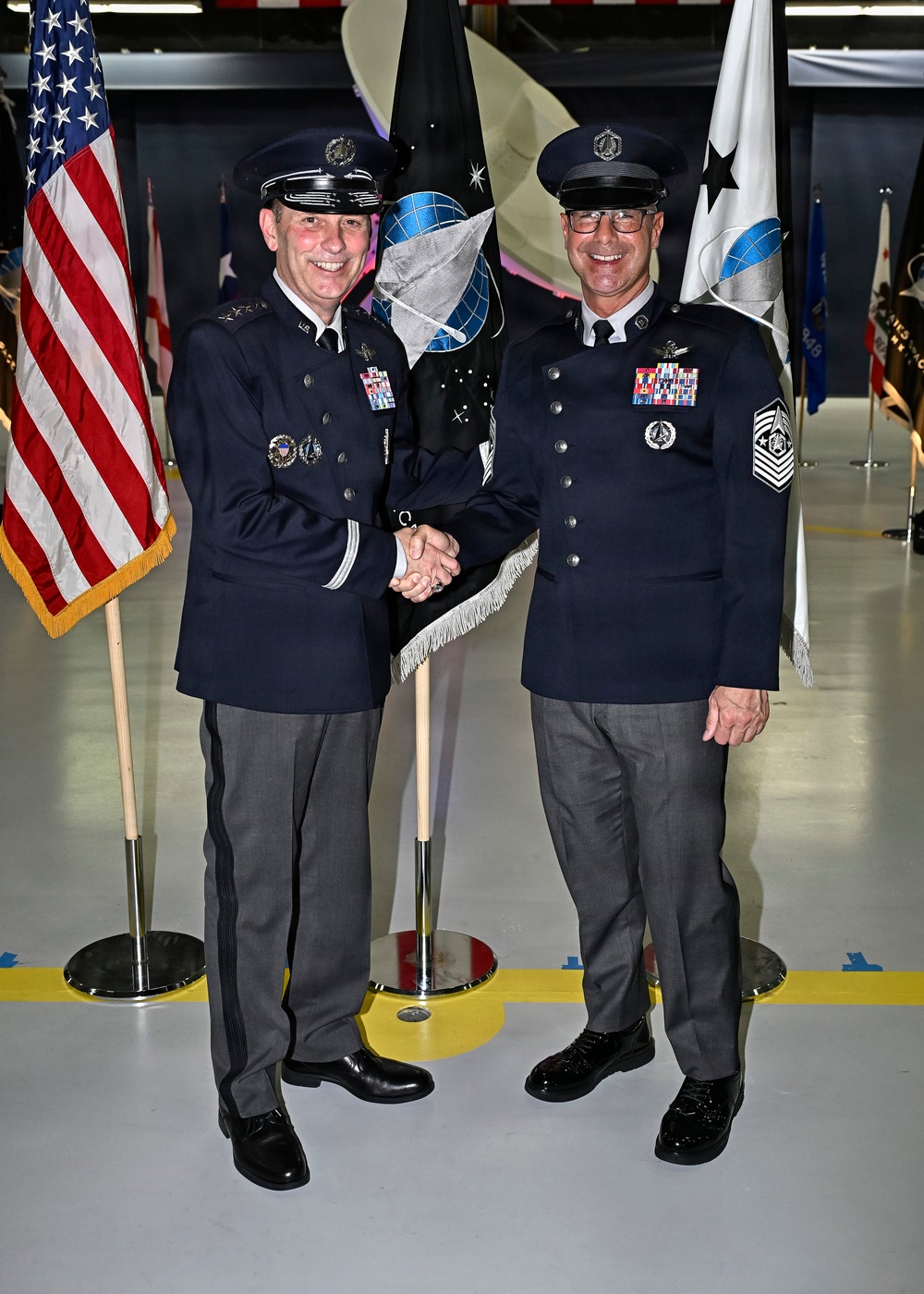 Two military personnel in dress uniforms are shaking hands and smiling during what appears to be a formal military ceremony. The individual on the left displays general officer rank insignia (three stars visible on shoulder boards), while the individual on the right wears Chief Master Sergeant rank insignia with Space Force or Air Force emblems. The setting is an indoor venue, likely a hangar, decorated with multiple military flags including the U.S. flag and unit guidons.