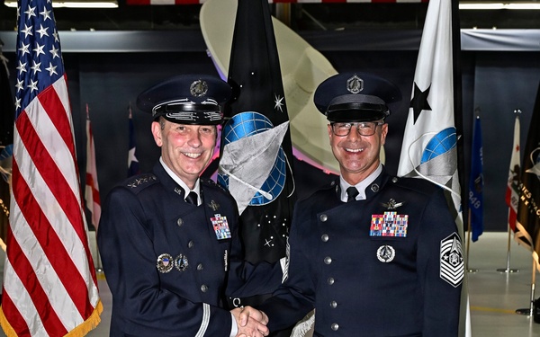 Two military personnel in dress uniforms are shaking hands and smiling during what appears to be a formal military ceremony. The individual on the left displays general officer rank insignia (three stars visible on shoulder boards), while the individual on the right wears Chief Master Sergeant rank insignia with Space Force or Air Force emblems. The setting is an indoor venue, likely a hangar, decorated with multiple military flags including the U.S. flag and unit guidons.