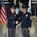 Two military personnel in dress uniforms are shaking hands and smiling during what appears to be a formal military ceremony. The individual on the left displays general officer rank insignia (three stars visible on shoulder boards), while the individual on the right wears Chief Master Sergeant rank insignia with Space Force or Air Force emblems. The setting is an indoor venue, likely a hangar, decorated with multiple military flags including the U.S. flag and unit guidons.