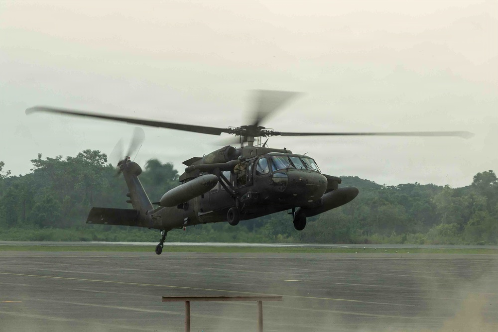 A military helicopter, consistent with a UH-60 Black Hawk, is captured in low hover or takeoff/landing phase above a wet tarmac runway. The aircraft is painted in olive drab/dark green military livery and appears to have external fuel tanks or equipment pods mounted on its sides. The setting appears to be a military airfield surrounded by dense tropical or subtropical vegetation under overcast skies.