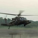 A military helicopter, consistent with a UH-60 Black Hawk, is captured in low hover or takeoff/landing phase above a wet tarmac runway. The aircraft is painted in olive drab/dark green military livery and appears to have external fuel tanks or equipment pods mounted on its sides. The setting appears to be a military airfield surrounded by dense tropical or subtropical vegetation under overcast skies.