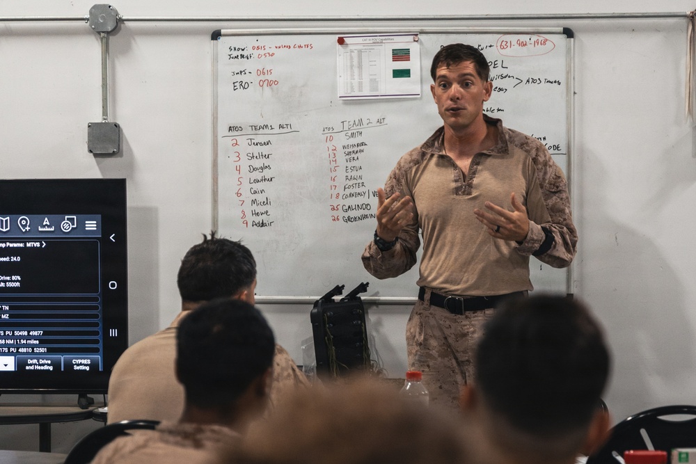 A military instructor in desert camouflage uniform is conducting a briefing or training session in front of a whiteboard. The whiteboard displays team rosters labeled 'ATOS TEAM 1' and 'ATOS TEAM 2' along with schedule times and other operational notes. Several personnel in military uniforms are seated facing the instructor.