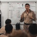 A military instructor in desert camouflage uniform is conducting a briefing or training session in front of a whiteboard. The whiteboard displays team rosters labeled 'ATOS TEAM 1' and 'ATOS TEAM 2' along with schedule times and other operational notes. Several personnel in military uniforms are seated facing the instructor.