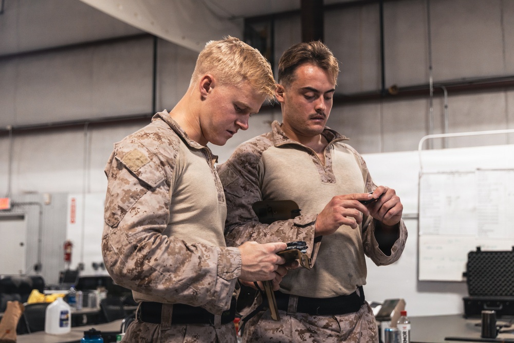 Two military personnel wearing USMC MARPAT desert camouflage uniforms are engaged in what appears to be weapons maintenance or inspection inside an indoor facility. Both individuals are focused downward, handling small components or tools. The setting appears to be a maintenance bay or armory workspace.