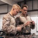 Two military personnel wearing USMC MARPAT desert camouflage uniforms are engaged in what appears to be weapons maintenance or inspection inside an indoor facility. Both individuals are focused downward, handling small components or tools. The setting appears to be a maintenance bay or armory workspace.