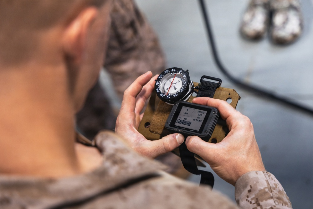 A military personnel in desert camouflage uniform is holding a combined navigation device consisting of a magnetic compass mounted on top of a small GPS/digital device with a tan MOLLE-style strap. The device displays a menu screen with options including 'DELETE MAP' and 'OPTIONS', along with coordinate data. The image is taken from an over-the-shoulder perspective near what appears to be a military vehicle.